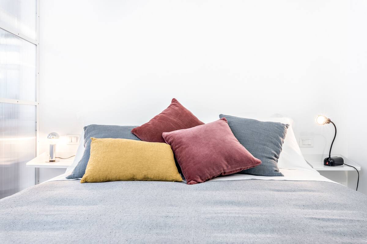 Ein ordentlich bezogenes Bett mit einer grauen Tagesdecke und drei Kissen in Rot, Gelb und Blau.