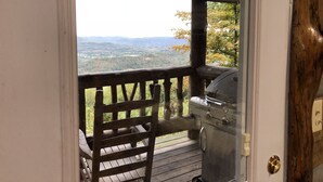 A wooden deck with a grill and a chair, overlooking a forested valley.