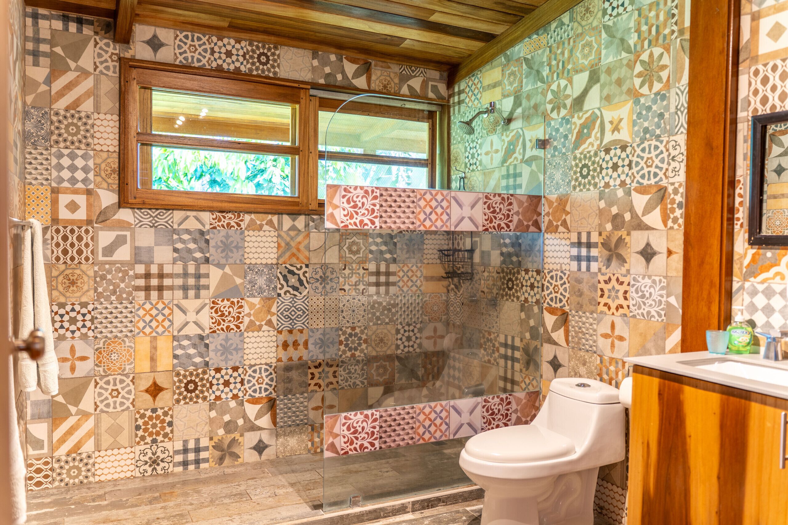 Une salle de bain avec différents types de carreaux décoratifs sur les murs et le sol, une fenêtre, des toilettes et un plafond en bois.