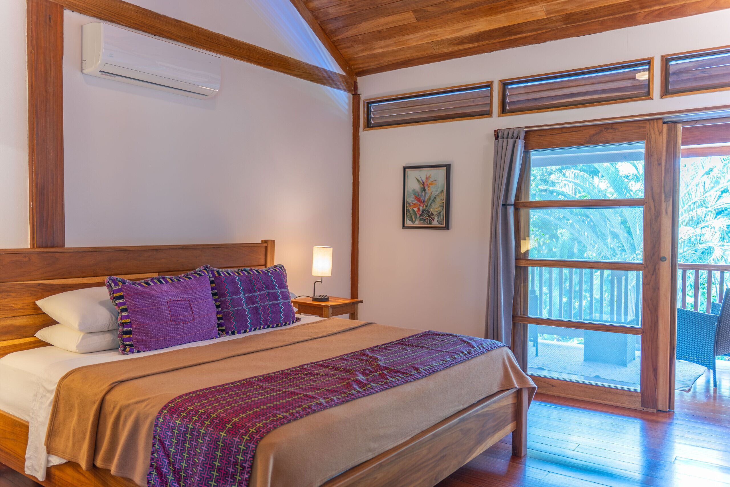 Una habitación con techo de madera, una cama con cabecera, una mesita de noche con lámpara, una ventana con vistas a la vegetación y un cuadro enmarcado en la pared.