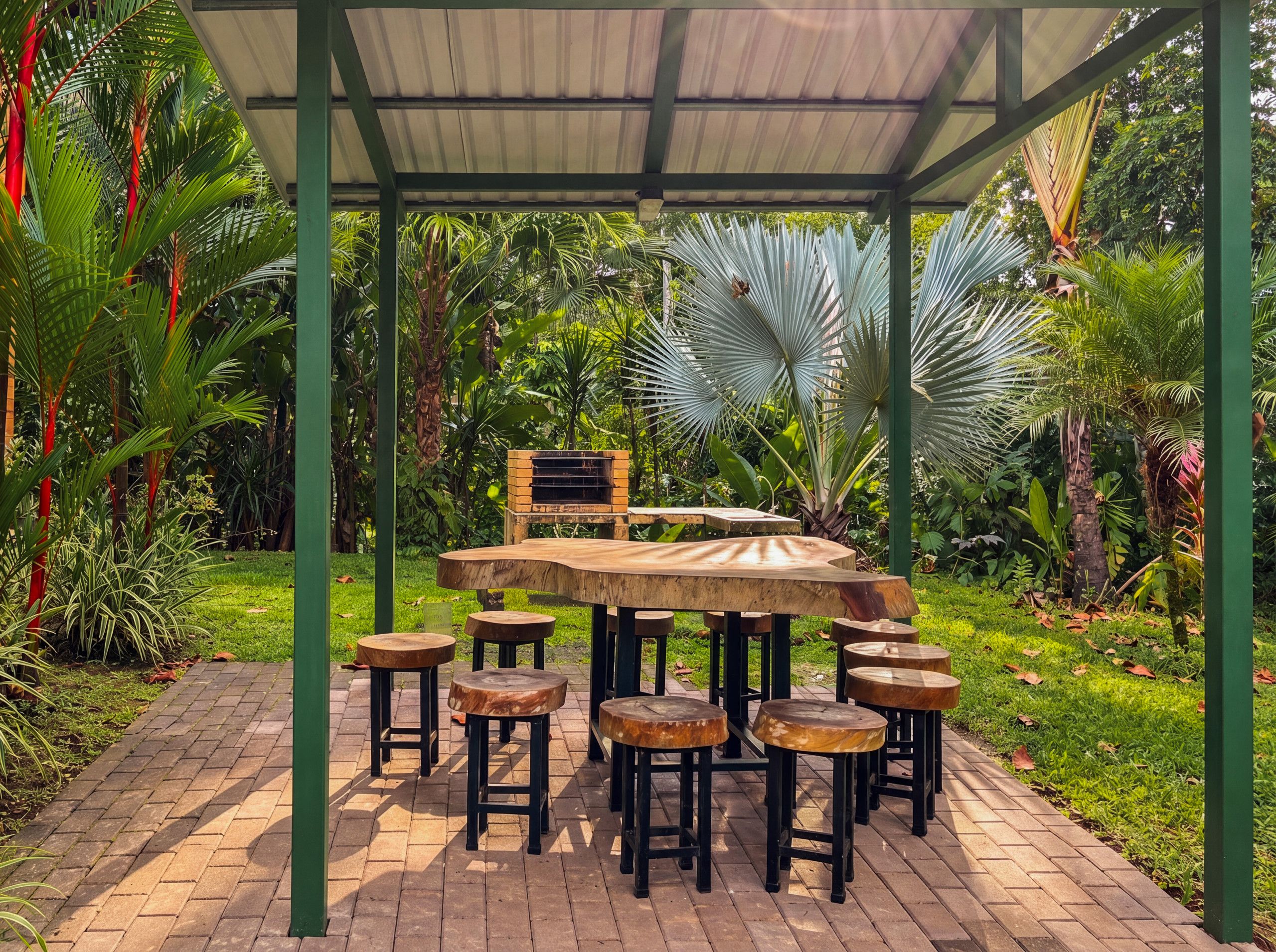 An outdoor patio with a wooden table and stools, surrounded by lush greenery and a metal roof structure.