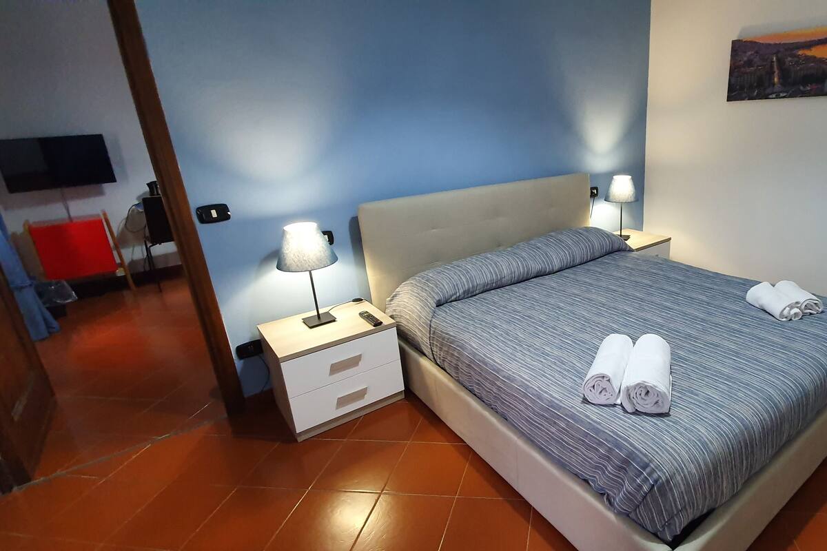 A neatly made bed with two rolled towels, a bedside lamp, and a flat-screen TV mounted on the wall.