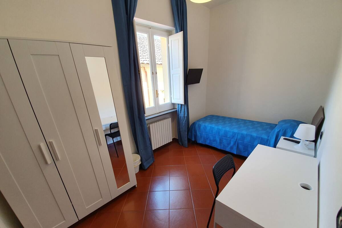 A small, tidy room with a bed, a desk, a chair, a wardrobe, and a window with curtains.