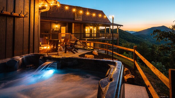 Een houten chalet met een jacuzzi en buiten zitplaatsen, met uitzicht op een bergachtig landschap.