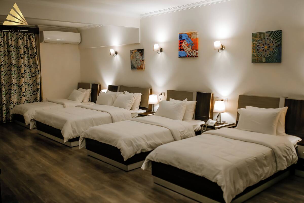 A hotel room with four beds arranged in a row, each with white bedding and nightstands with lamps.