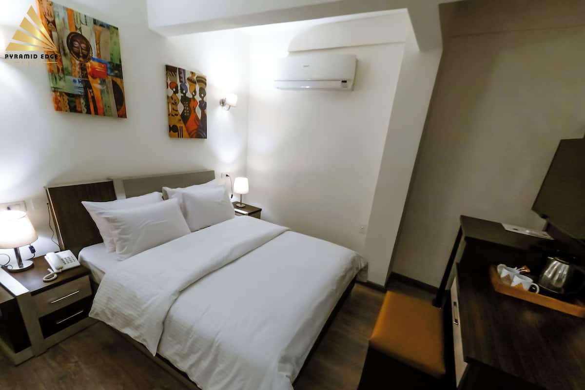 A hotel room with a bed, bedside tables, a lamp, an air conditioning unit, and a painting on the wall.