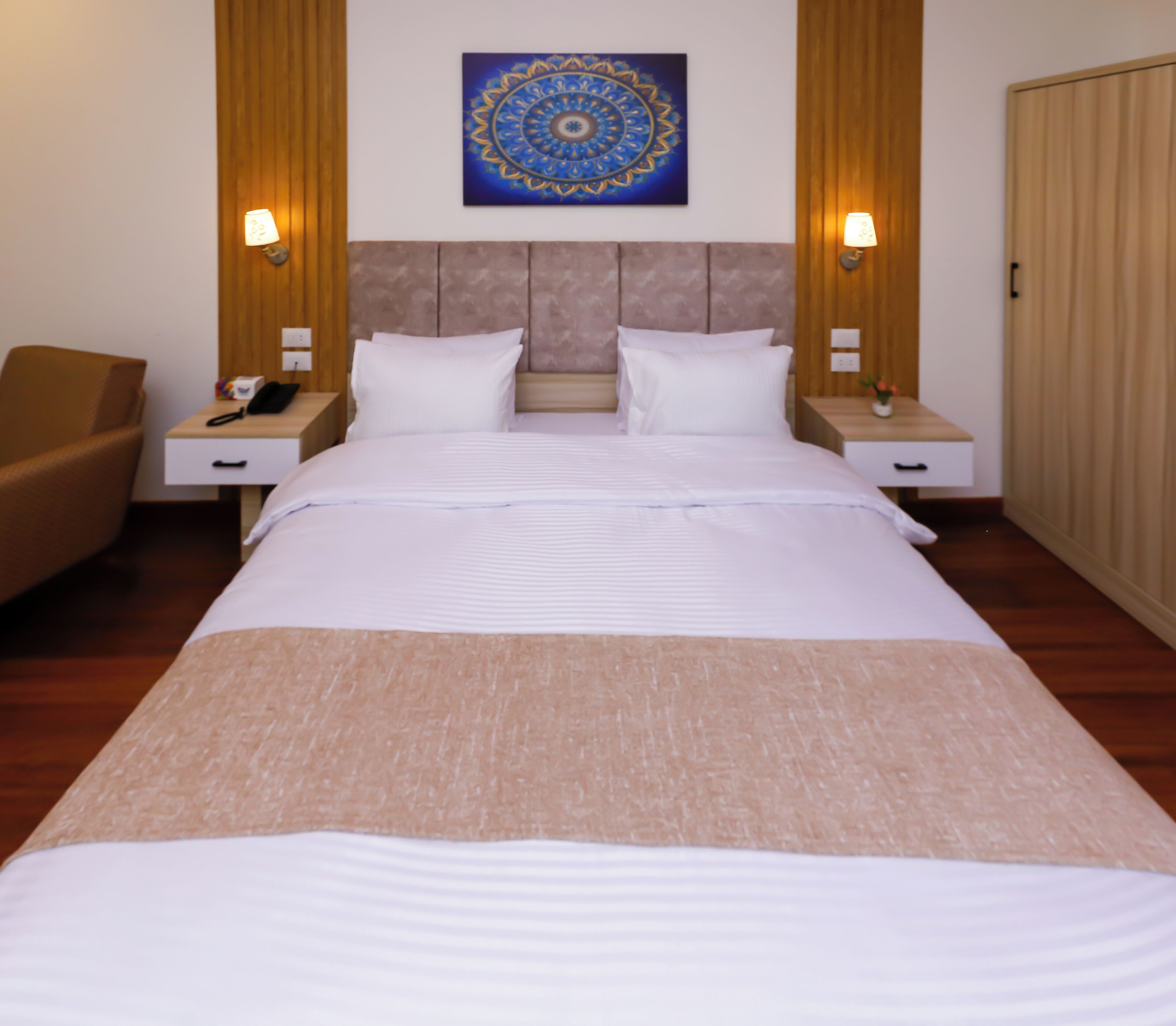 A hotel room with a large bed, two bedside tables, a chair, a wall-mounted artwork, and a wardrobe.