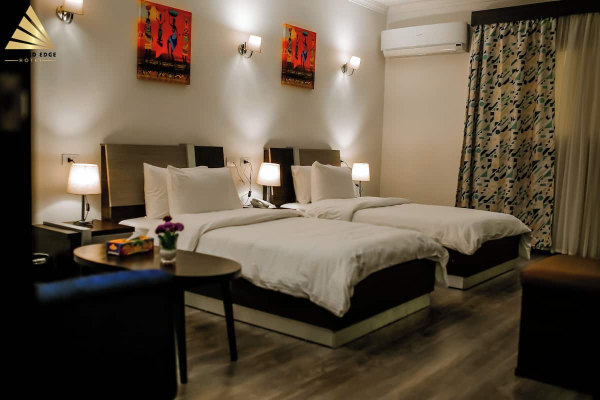 A hotel room with two beds, bedside lamps, a small table with a vase, and artwork on the walls.