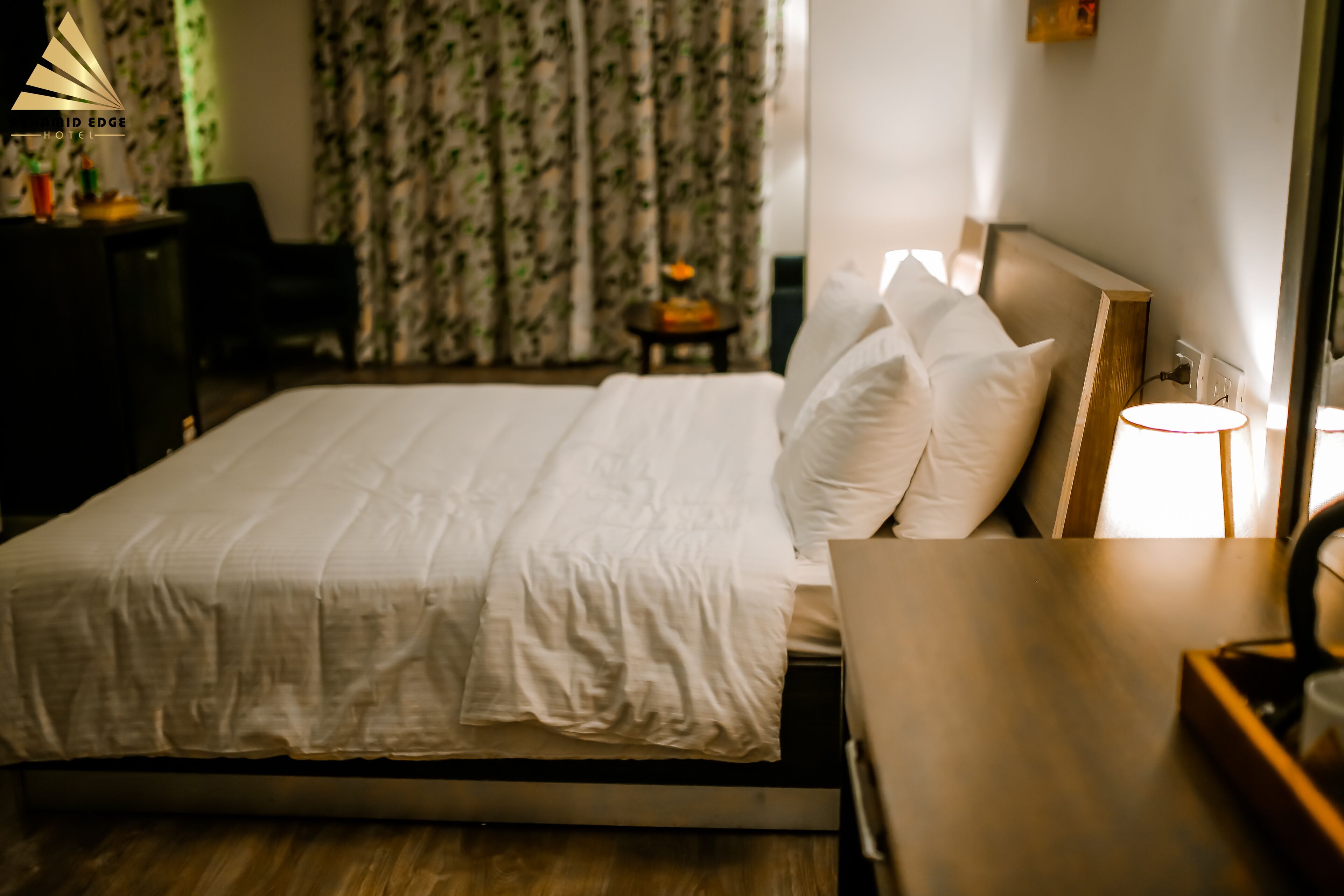 A neatly made bed with white linens and pillows, a bedside table with a lamp, and a small table with a fruit bowl in the background.