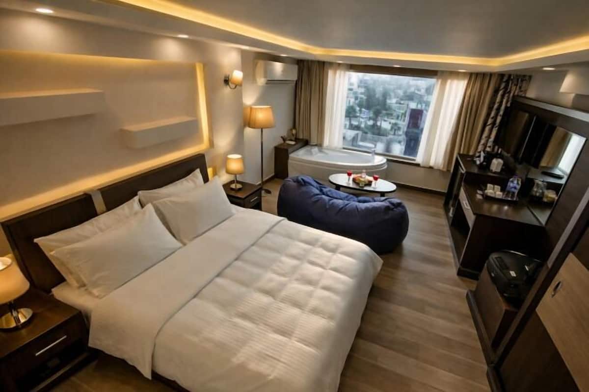A hotel room with a bed, a desk, a chair, a small table with a drink, and a window with a view.
