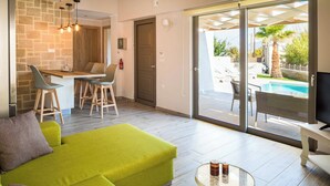 A modern living room with a green sofa, a glass coffee table, and a TV on a white stand. There is a dining area with a wooden table and chairs, and a large sliding glass door leading to an outdoor pool area. - Villa Elenitsa - Two Bedroom Villa, Sleeps 4 (Minoa Pediada)