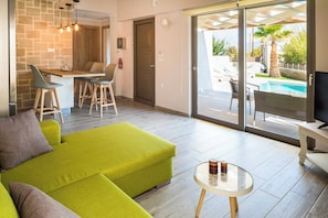 A modern living room with a green sofa, a glass coffee table, and a TV on a white stand. There is a dining area with a wooden table and chairs, and a large sliding glass door leading to an outdoor pool area.