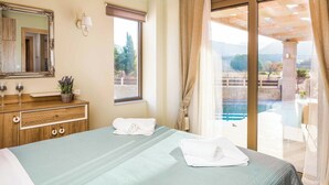 A hotel room with a bed, a wooden dresser, a mirror, a window with a view of a pool and mountains, and a sliding glass door to an outdoor area. - Villa Elenitsa - Two Bedroom Villa, Sleeps 4 (Minoa Pediada)
