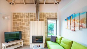 A living room with a stone wall, a fireplace, a green sofa, and a television on a white stand. - Villa Elenitsa - Two Bedroom Villa, Sleeps 4 (Minoa Pediada)