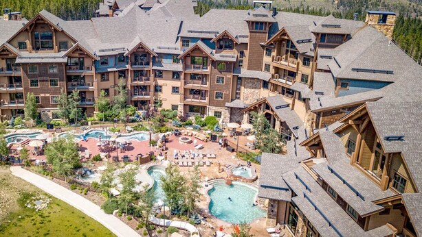 Un resort con diversi edifici, una piscina centrale circondata da lettini prendisole e montagne sullo sfondo.