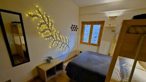 Et soveværelse med køjeseng, et natbord med en plante, en vægmonteret lampe og et vindue med udsigt til træer.