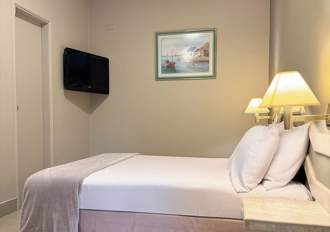 A hotel room with a bed, a flat-screen TV mounted on the wall, a painting above the bed, and a bedside lamp.