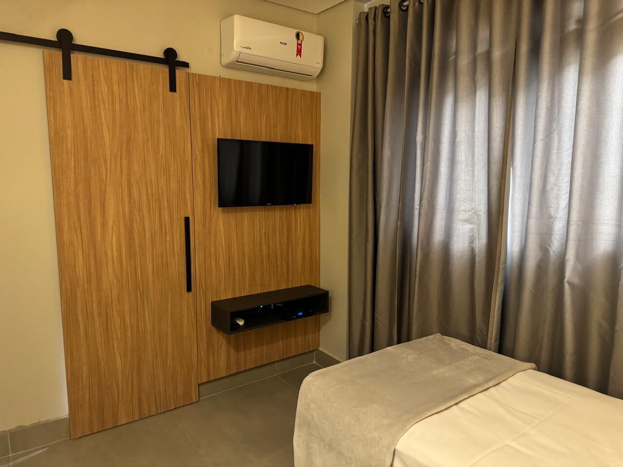 A hotel room with a wooden wardrobe, a mounted flat-screen TV, a bedside table, and a bed with a white cover.
