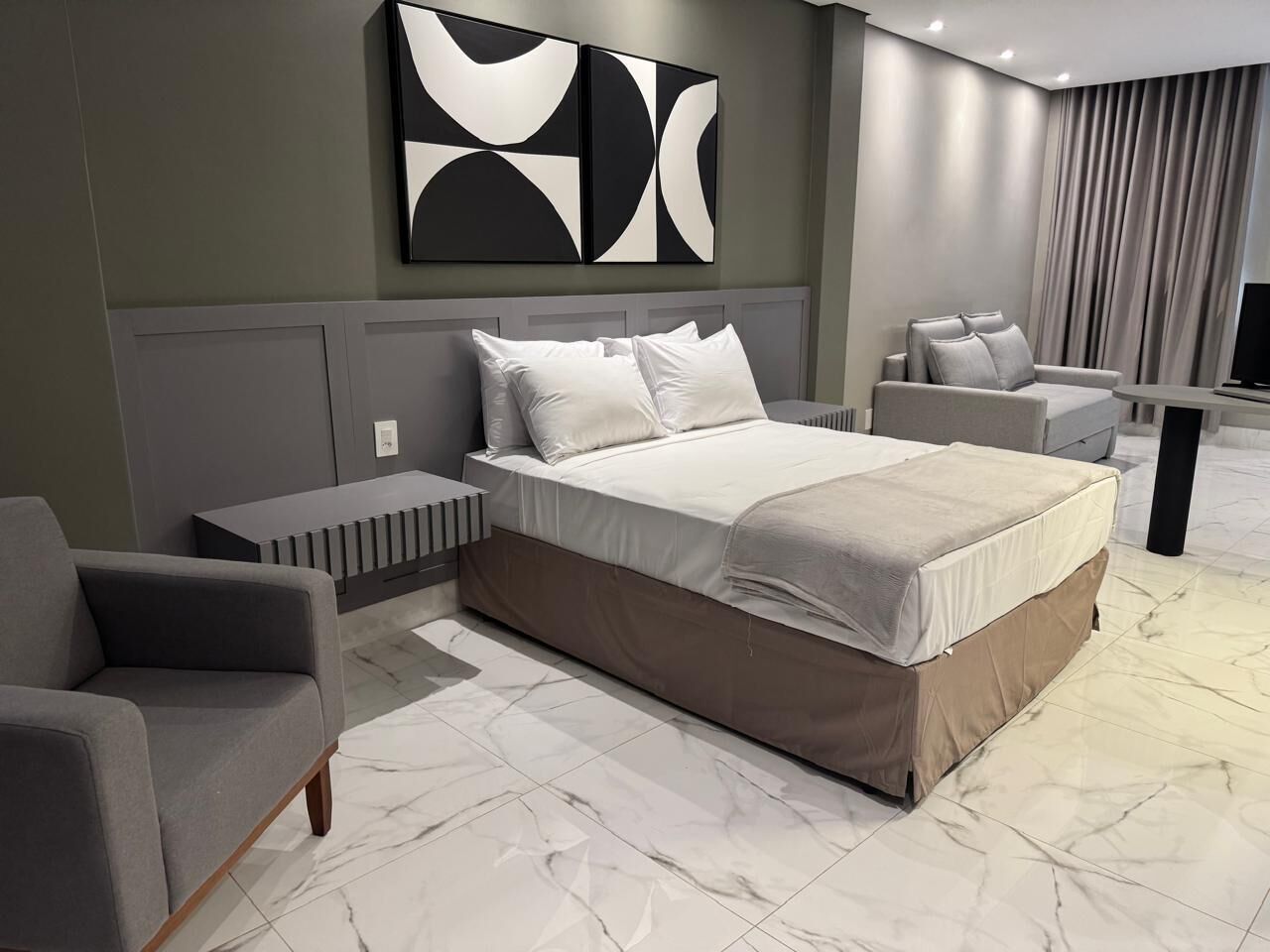 Un dormitorio moderno con una cama grande, un sofá gris, una mesita pequeña y un televisor.