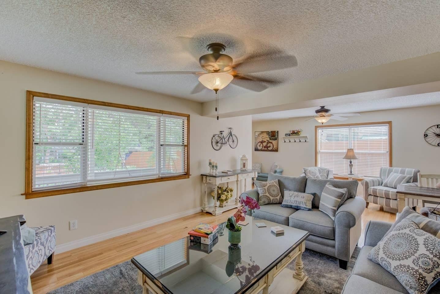 A spacious living room with a large window, a ceiling fan, a sofa set, and a coffee table.