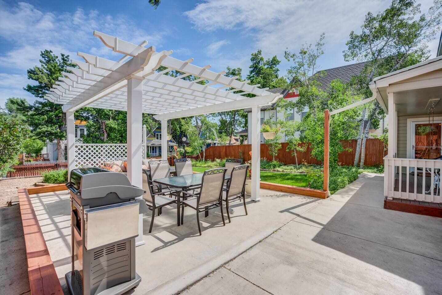 Un patio con una parrilla, mesa de comedor y sillas bajo una pérgola blanca.