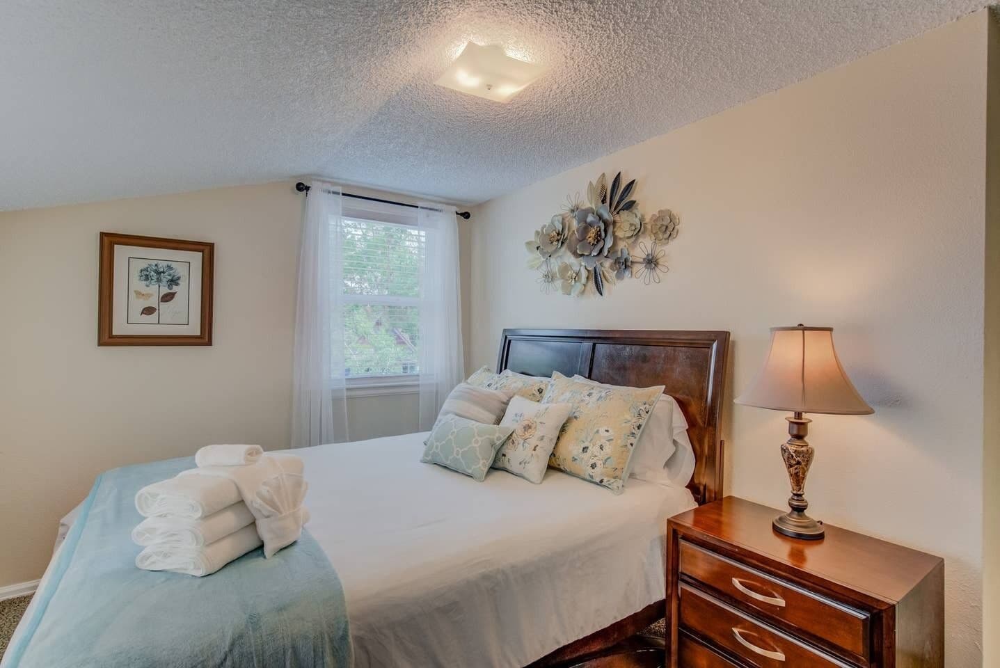 A neatly made bed with a blue blanket and white towels, a wooden bedside table with a lamp, a window with sheer curtains, and a wall-mounted floral decoration.