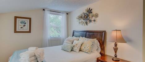 A neatly made bed with a blue blanket and white towels, a wooden bedside table with a lamp, a window with sheer curtains, and a wall-mounted floral decoration.