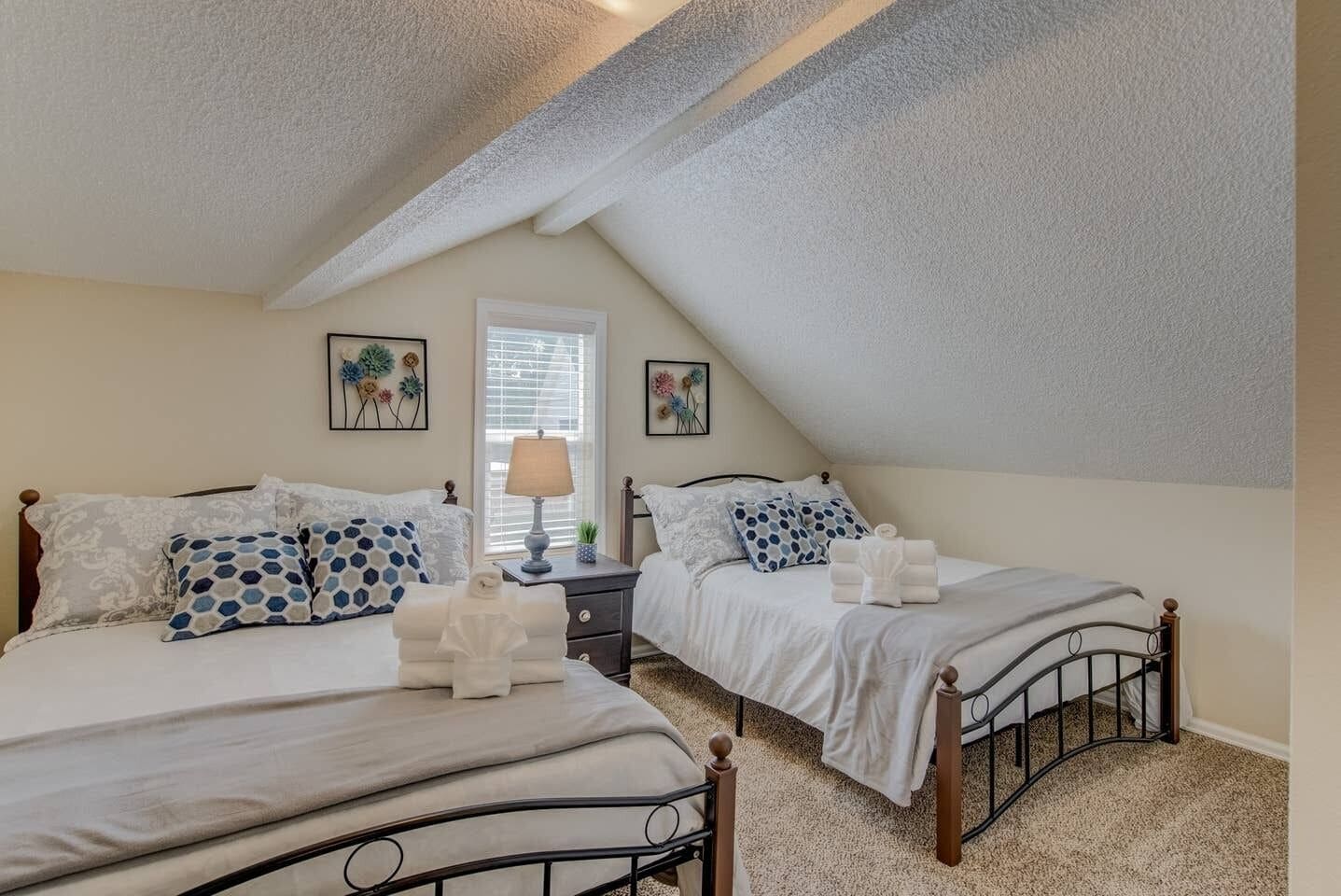 A bedroom with two beds, a window with blinds, and a ceiling with a sloped design.