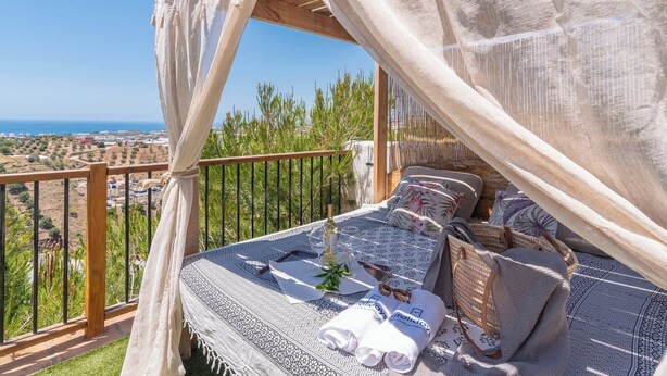 A balcony with a bed, a table with a glass, and a view of the landscape.