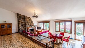 Uma sala de estar ampla com lareira de pedra, um armário de madeira, uma televisão e um espaço de refeições com cadeiras vermelhas.