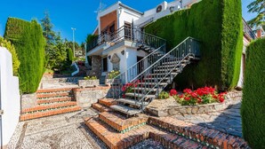 Uma casa de dois andares com uma escadaria que dá para uma varanda, cercada por sebes bem cuidadas e um jardim com flores vermelhas.
