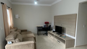 Un salon moderne avec un ensemble de canapés beige, un téléviseur à écran plat posé sur un meuble en bois, un bureau avec un ordinateur et un ventilateur de plafond.
