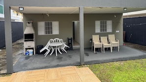 Une terrasse extérieure couverte, équipée d’un barbecue, d’une table avec des chaises et d’une pile de chaises supplémentaires.