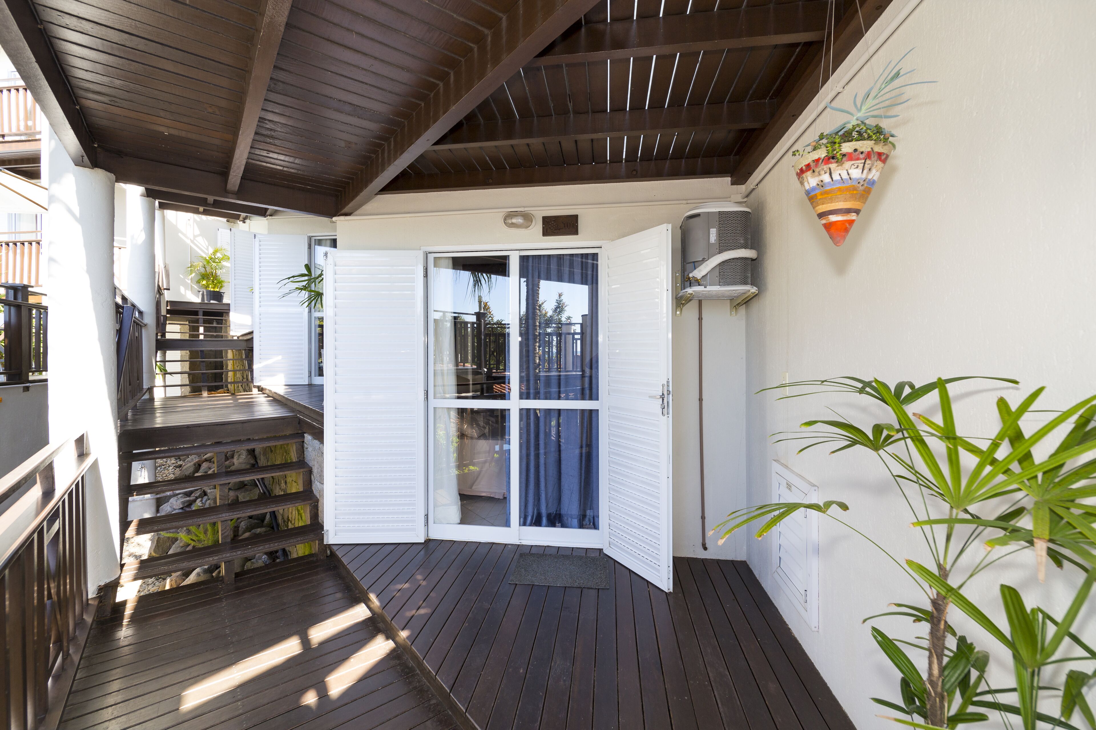 A covered outdoor area with a wooden deck, a set of stairs leading to a glass door, and a potted plant.