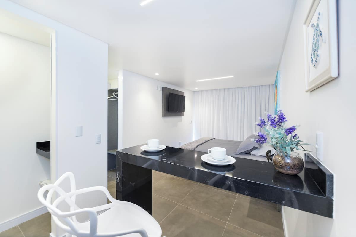 Une chambre d’hôtel moderne avec un comptoir en marbre noir, des chaises blanches, un téléviseur à écran plat fixé au mur et un vase avec des fleurs violettes posé sur le comptoir.
