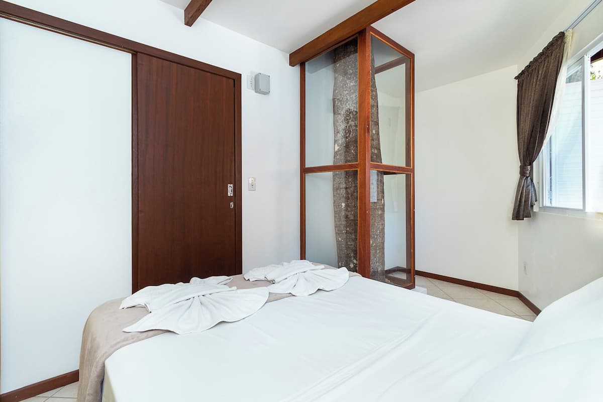 Une chambre avec une porte en bois, une cloison en verre, un lit avec du linge de lit blanc et une fenêtre avec des rideaux.
