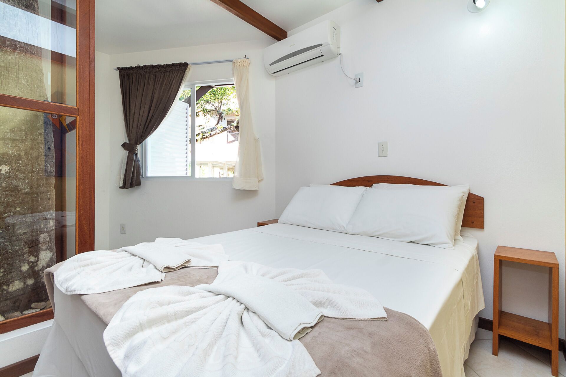 Une chambre à coucher avec un lit, une fenêtre avec des rideaux, un appareil de climatisation et une petite table de chevet en bois.