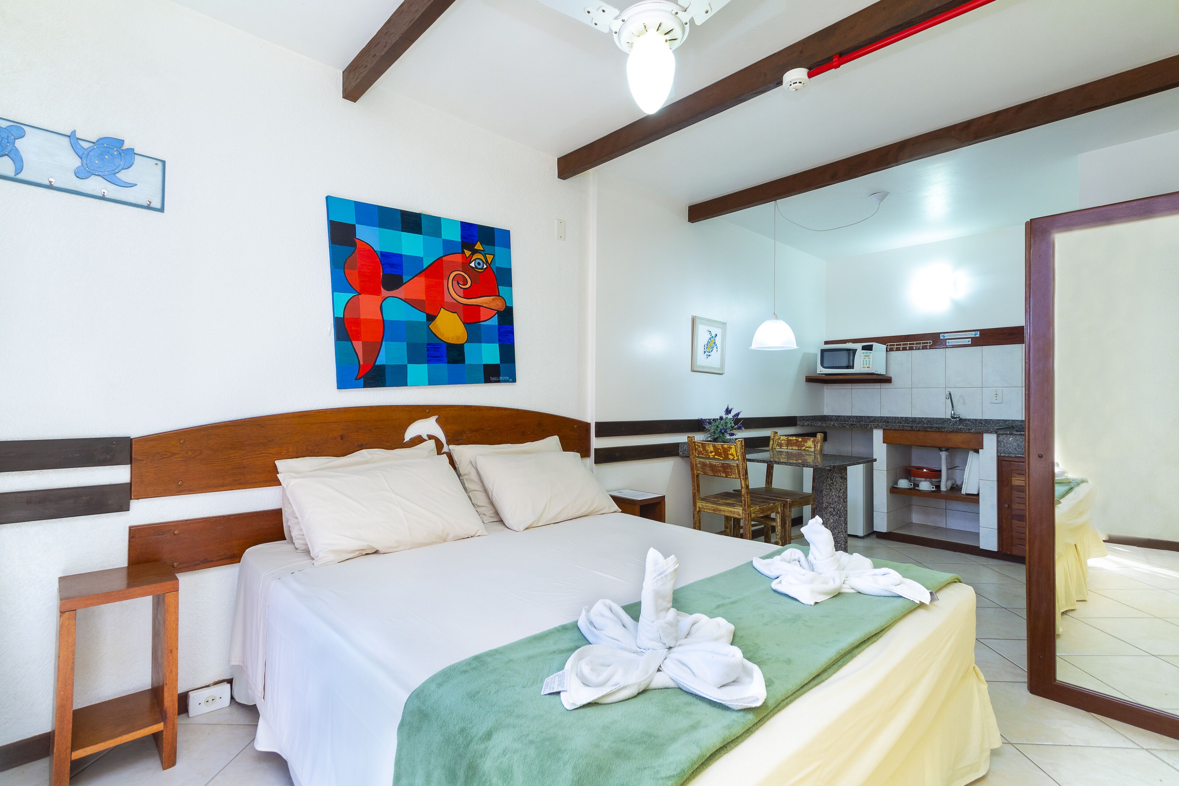 A hotel room with a bed, a wooden headboard, a painting, a green towel, and a view of the kitchen.