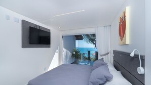 A modern bedroom with a large bed, a flat-screen TV mounted on the wall, a ceiling light, and a view of the ocean through a sliding door. - pousada solar das tartarugas (Bombinhas)