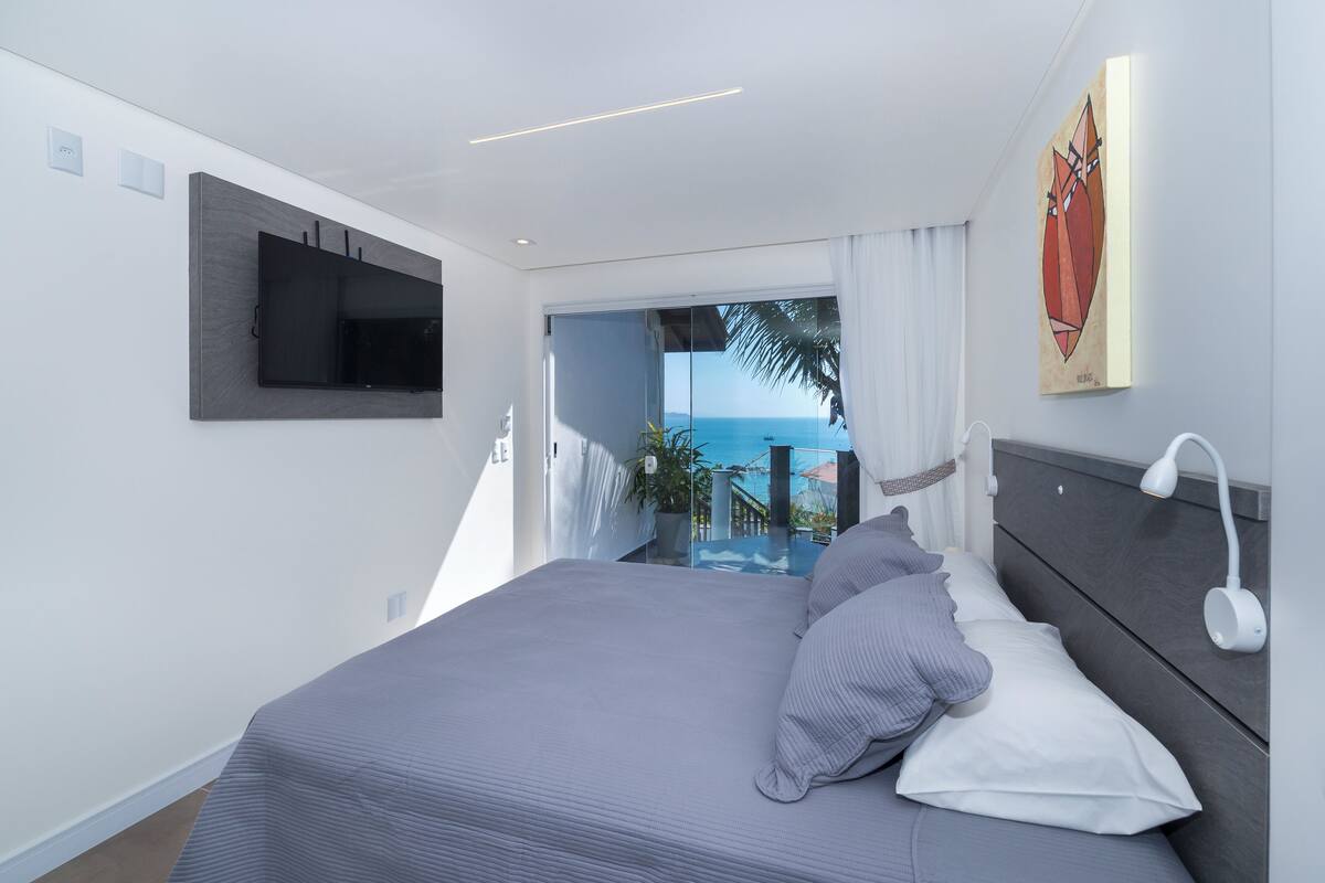 Une chambre moderne avec un grand lit, un téléviseur à écran plat fixé au mur, un luminaire au plafond et une vue sur l’océan par une porte coulissante.