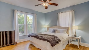 A bedroom with a bed, a ceiling fan, a window with curtains, a bedside table with a lamp, and a radiator.