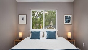 A bedroom with a large bed, two bedside tables with lamps, a window with a view of trees, and two framed pictures on the walls.