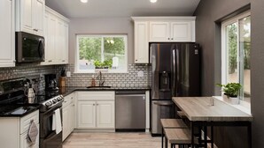 A modern kitchen with white cabinetry, a stainless steel refrigerator, a coffee machine, and a window with a view of greenery.