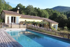A pool area with a wooden deck, a fenced garden, and a building with a tiled roof. - House with heated pool in the Val d´Emeraude (lasalle)