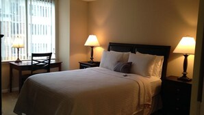 A neatly made bed with a white comforter and pillows, two bedside lamps, a chair, and a window with blinds. - National at Ballston Metro (Arlington)