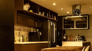 A modern kitchen with dark cabinetry, a stainless steel refrigerator, and a wooden dining table. - Apto de luxo em Jurerê internacional REN002 (Florianópolis)