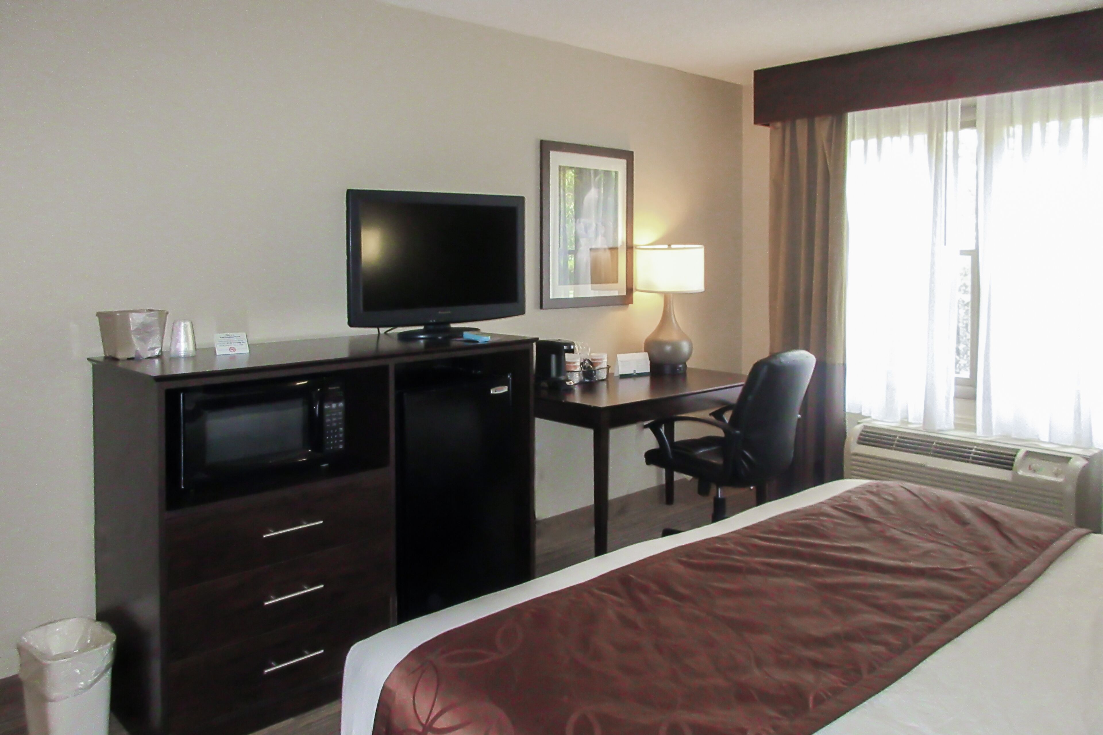 Quarto de hotel com cama, escrivaninha com computador, televisão, micro-ondas e lixeira.
