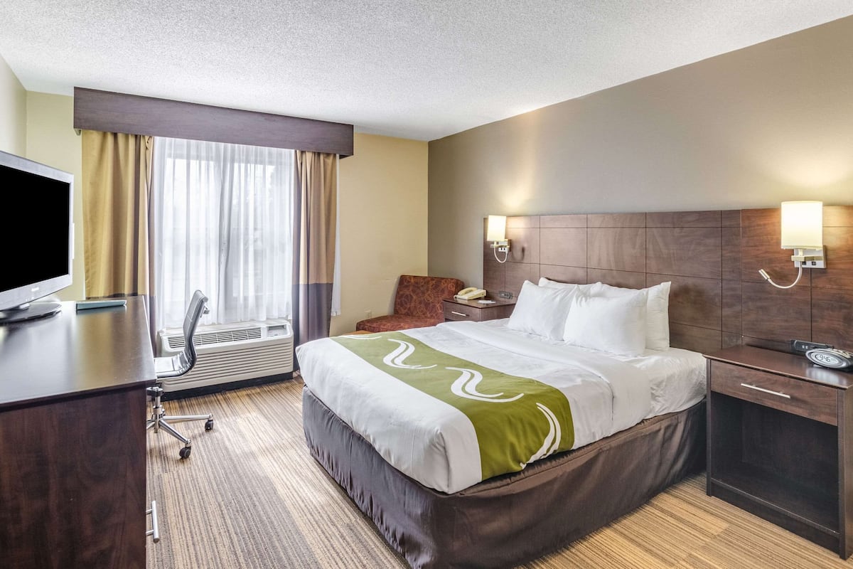 Ein Hotelzimmer mit Bett, Schreibtisch, Stuhl, Nachttisch, Lampe und Fernseher.