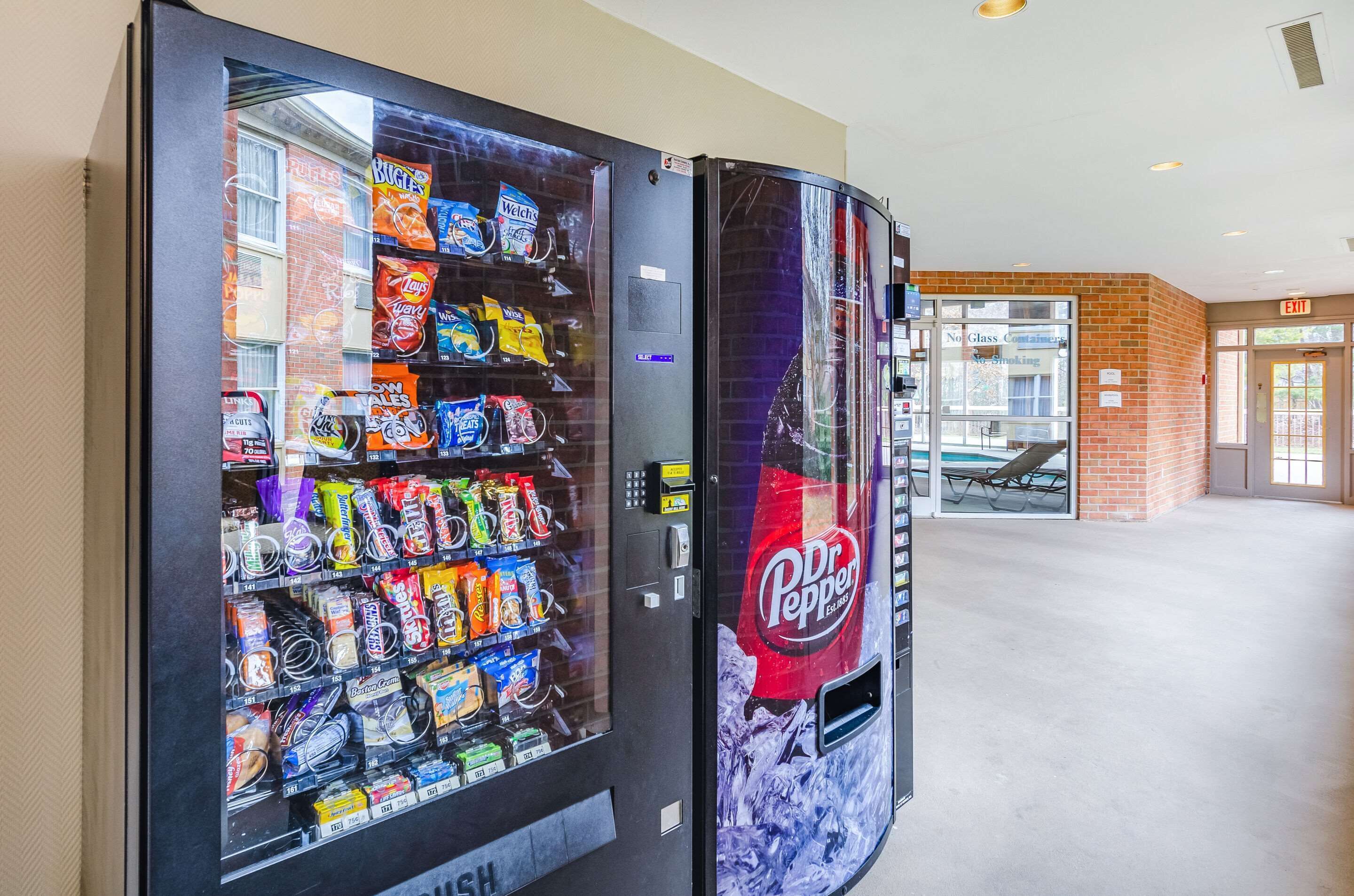 A vending machine stocked with snacks and beverages, including Dr. Pepper, in a hallway with a brick wall and an exit sign.