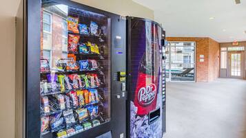A vending machine stocked with snacks and beverages, including Dr. Pepper, in a hallway with a brick wall and an exit sign.